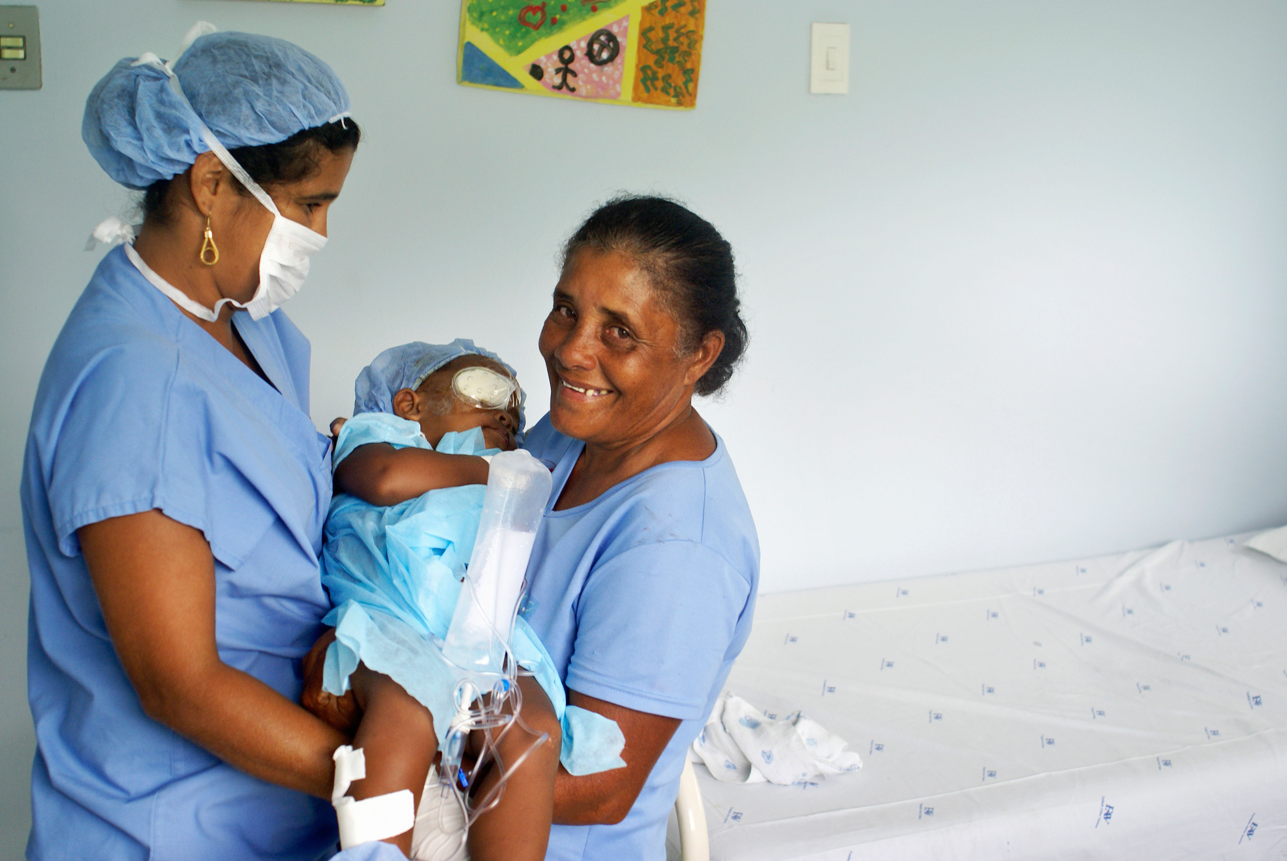 Two midwives holding a baby with an eye patch after surgery