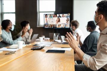 People wearing masks sit around a table and wave to online participants during a webinar